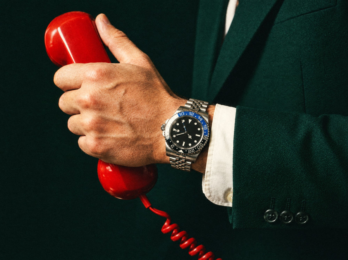 Mano con un reloj GMT de bisel azul y negro, tomando un teléfono rojo